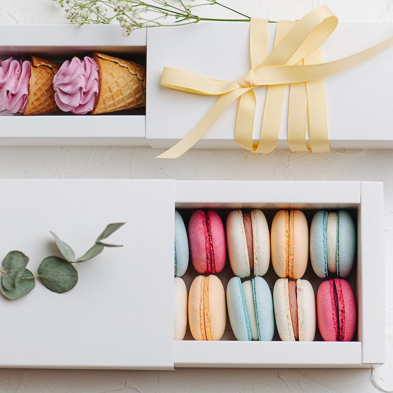 Two rectangle pull out boxes with sweets parallel to each other. Colorful macarons and waffle cones with zefir on top. On a white surface. Arranged and decorated for composition.
